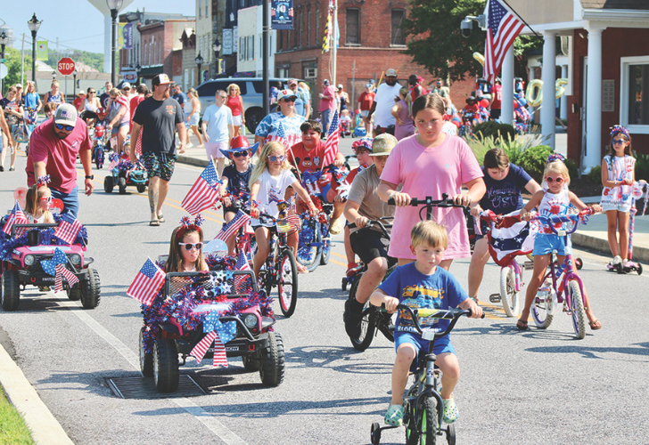 Kids Parade 1 Children riding bikes and toy vehicles along a sunny street parade, many bearing American flags, with spectators in the background.