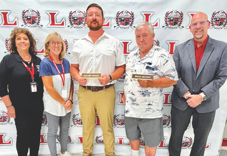 Laurel Board 4 Five adults pose for a photo in front of an event backdrop with logos; the central man holds a plaque labeled 'ZACHARY WHALEY' while a man next to him holds a name badge 'BRENT NICHOLS'.