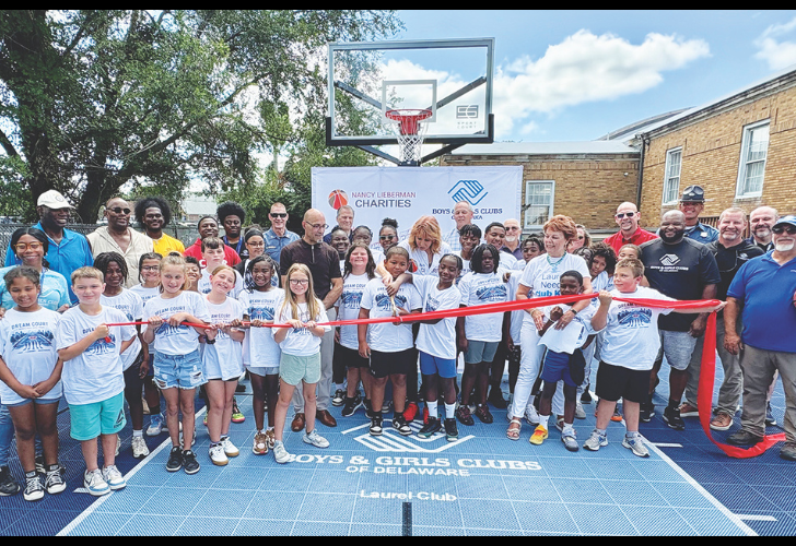Group of children and adults at a ribbon-cutting on a blue basketball court for a Boys & Girls Clubs event, with Nancy Lieberman Charities banner in the background.