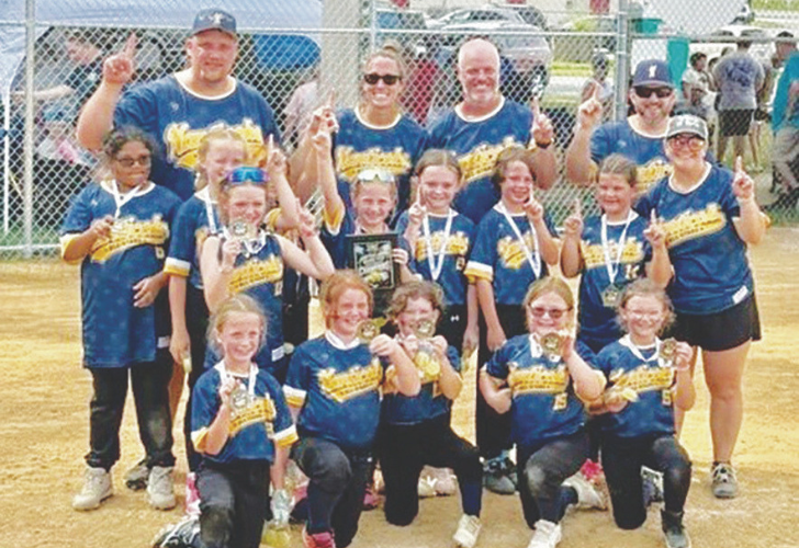 Machine pitch- Seaford 30 Young girls in blue team jerseys with medals pose on a softball field, holding a trophy with coaches behind them.