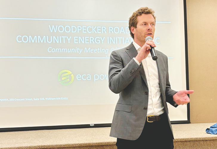 Man in a gray suit speaks into a handheld microphone at a community energy meeting, with a slide projection in the background.
