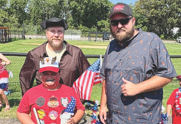 Maryland 4 Three people pose outdoors at a patriotic event: a boy in a red shirt with helmet graphics holds a small cup and flag while two men stand beside him, one giving a thumbs up.