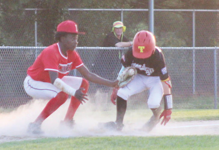 STOLEN BASE 42 Two young baseball players collide at a base: one in red slides while the other in black with a yellow helmet guards the bag, dust rising as a spectator watches behind a chain-link fence.