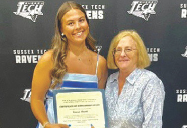 Scholarships2 6 Young woman in a blue dress holds a certificate while standing with an older woman in a light patterned shirt at a school awards backdrop.