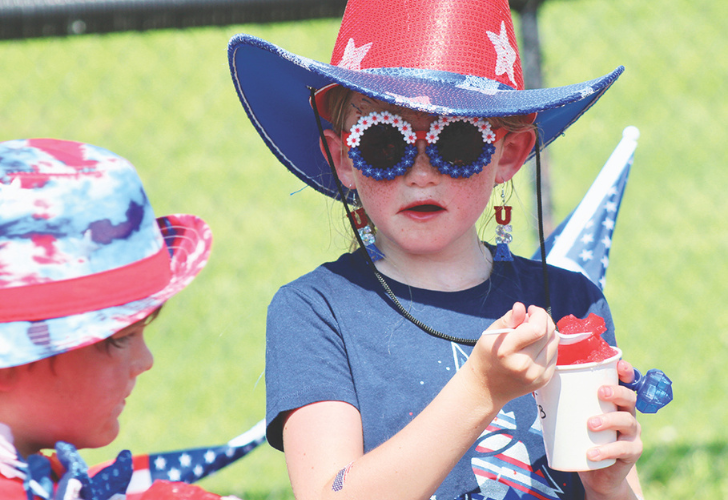 Two children in patriotic outfits outdoors, wearing red‑white‑blue hats and star sunglasses; one holds a white cup with a red treat.