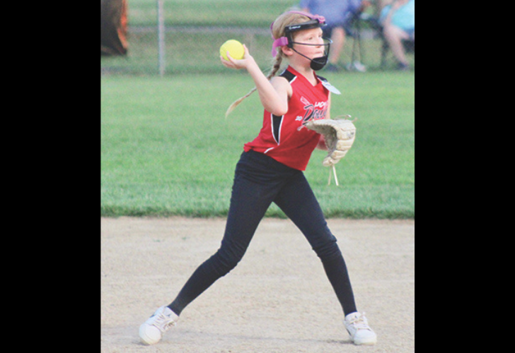 THROW TO FIRST2 40 Young girl in a red softball uniform pitches a ball on a dirt infield, wearing a helmet and glove.