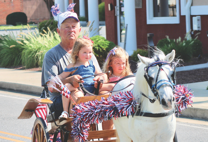 Vehicles 4 Grandfather with two young girls riding a festive pony-drawn wagon in a town parade, decorated in red, white, and blue garlands.