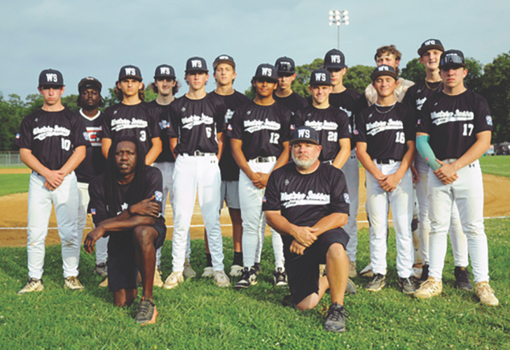 Baseball team in black uniforms posing for a group photo on the infield grass, with two coaches kneeling in front.