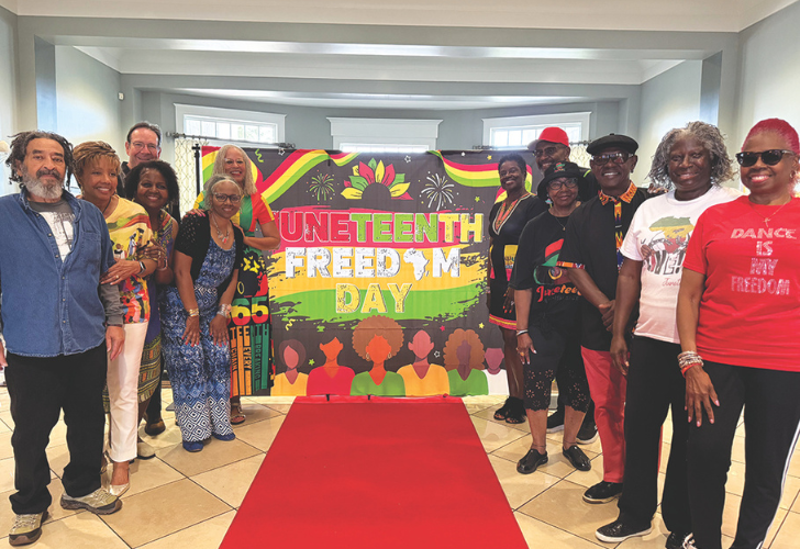 A diverse group of adults posing indoors beside a bright Juneteenth Freedom Day banner on a red carpet.