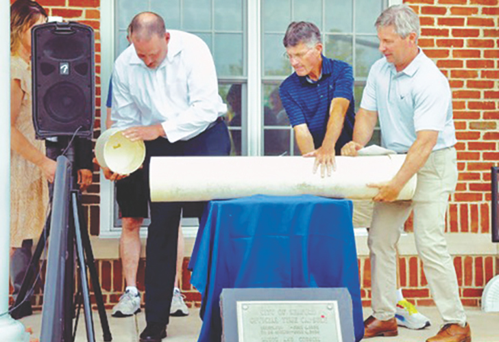 time capsule People at a dedication ceremony unrolling a large blueprint in front of a brick building outside a doorway.