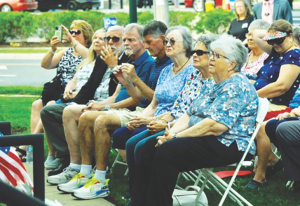 Audience of mostly seniors seated on folding chairs at an outdoor event; several wear sunglasses, one person photographs with a phone, an American flag is visible in the foreground.